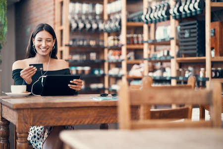 Online shopping with a credit card at a cafe by a trendy woman browsing on a tablet with earphones and paying for her bill at a coffee shop. Happy woman enjoying a break and downloading a finance appの写真素材