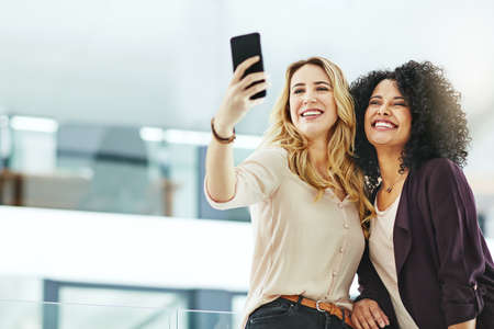 Happy, smiling and cheerful women taking selfies with a phone while bonding, having fun and being carefree at work. Diverse, joyful and positive colleagues taking photos for social mediaの写真素材