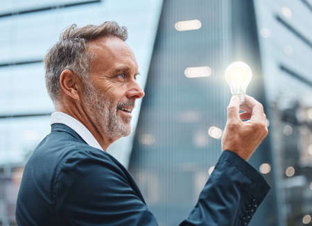 The best tool in a business kit is a bright mind. a mature businessman holding a lightbulb against an urban background.の写真素材