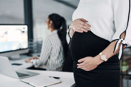 Motherhood wont stop me reaching goals. an unrecognizable pregnant businesswoman standing and holding her stomach while a colleague works behind her.の写真素材