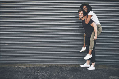 Fun, trendy and happy interracial couple enjoying a piggyback ride together outside against an urban background. Lovers bonding and being playful. Smiling and relaxing while walking outdoorsの写真素材