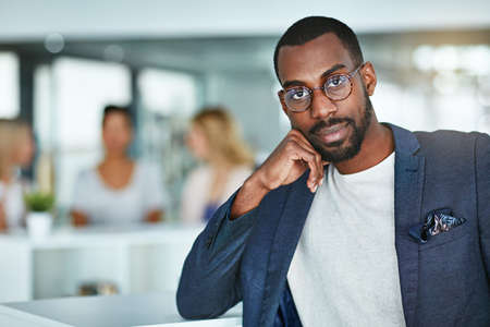 Portrait of a confident black business man standing in a busy office. Proud, stylish and ambitious professional focused on his success and career while his employees have a meeting in the backgroundの写真素材