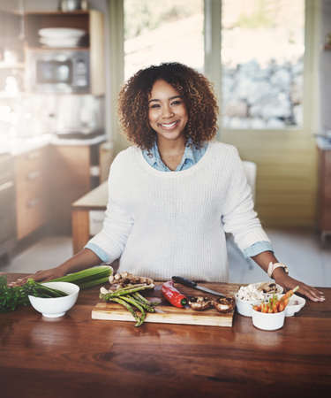 Portrait of happy black woman preparing healthy food in a kitchen at home. Young African American using fresh vegetables to make a delicious, balanced low carb meal. Lady on a cleanse and detox dietの写真素材