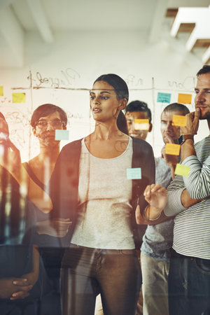Group of marketing professionals or designers brainstorming ideas, writing on transparent board with sticky notes during meeting. Diverse team of colleagues discussing work project schedule plan.の写真素材
