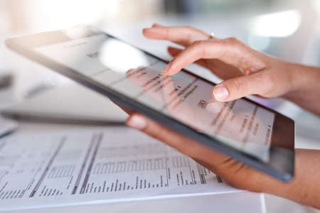 Lets see whats on the menu for my workday. Closeup shot of an unrecognisable businesswoman using a digital tablet while going through paperwork in an office.の写真素材