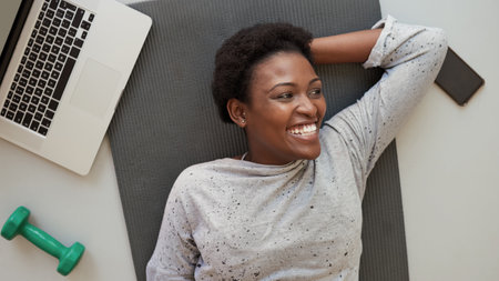 Theres a natural symbiosis between fitness and personal development. High angle shot of a young woman lying on an exercise mat with a laptop, cellphone and dumbbell around her.の写真素材
