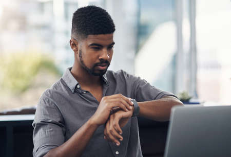 Meeting deadlines right on time. a young businessman checking the time while working on a laptop in an office.の写真素材