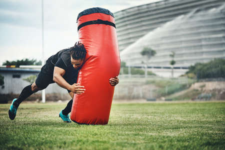 Train today, win tomorrow. Full length shot of a handsome young rugby player working out with a tackle bag on the playing field.の写真素材