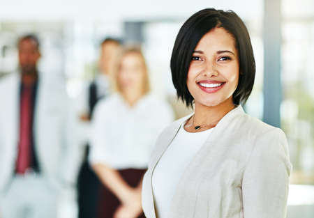 Young, smart and smiling businesswoman with a diverse team at work ready to achieve success. Confident female professional executive standing in a modern office with her colleagues in the backgroundの写真素材