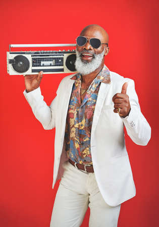 Music is what uplifts the spirit. Studio shot of a senior man wearing vintage clothing while posing with a boombox against a red background.の写真素材