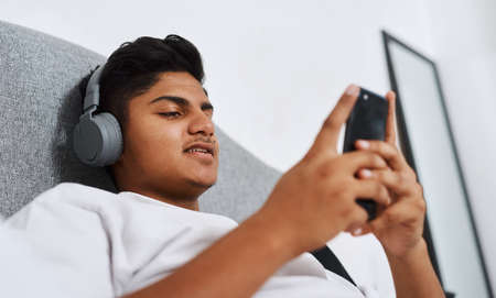Streaming some new releases. a young man using his cellphone while listening to music through his headphones.の写真素材