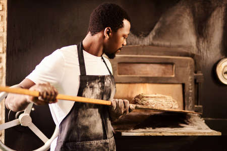 I make them fresh every day. a male baker removing freshly baked bread from the oven.の写真素材