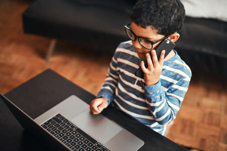 Hes a busy little man. an adorable little boy talking on a cellphone while using a laptop at home.の写真素材