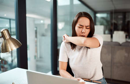 Practice good cough etiquette for the sake of everyones health. a young businesswoman coughing into her elbow in an office.の写真素材