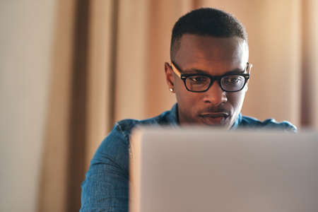 Focused, serious and confident entrepreneur typing on a laptop and reading an email sitting alone in his home office. African american businessman planning and remote working his startup businessの写真素材