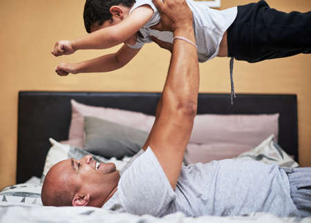 a cheerful young man lifting up his son in the air to fly like a super hero while lying down on a...
