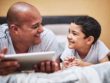 That was a fun episode. a cheerful young man and his son watching videos on a digital tablet together while hanging out on a bed at home during the day.の写真素材