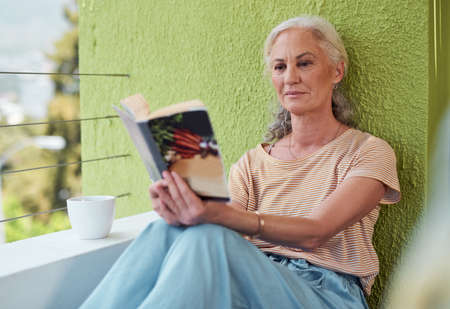 Doing retirement right. a mature woman reading a book and having coffee on her balcony at home.