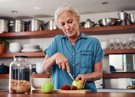 These days, she only eats healthy. an attractive senior woman chopping up apples and other fruit while preparing breakfast in the kitchen.の写真素材