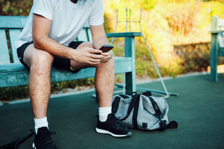 Connectivity on the courts. Closeup shot of an unrecognisable man using a cellphone while sitting on a bench on a tennis court.の写真素材