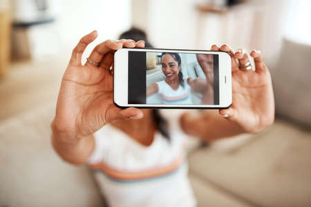 Living life online. Cropped portrait of an attractive young blogger sitting in her home and using her cellphone to take a selfie.の写真素材
