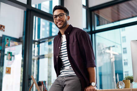 Lets get this day started. Cropped portrait of a handsome young businessman wearing spectacles and leaning on his desk while working alone in his office.の写真素材