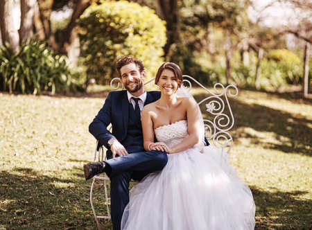 Were so happy we finally tied the knot. Cropped portrait of an affectionate newlywed couple smiling while sitting outdoors on their wedding day.の写真素材