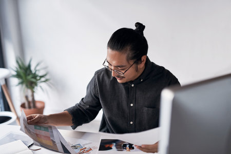 So much paperwork to go through. a handsome young businessman looking at paperwork and sitting alone at his desk in the office.の写真素材
