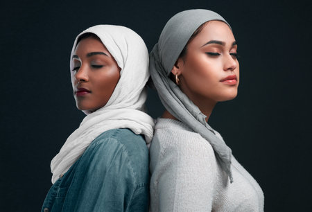 And where she stood, she stood tall. two attractive young women wearing hijabs and standing back to back against a black studio background.の写真素材