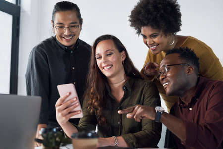 Technology has made business on the go more convenient. a diverse group of businesspeople looking at a cellphone together in a modern office.の写真素材