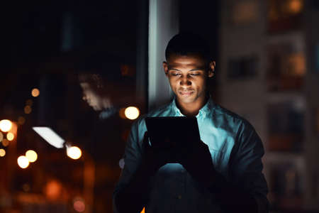 Quickly touching base with the business world. a handsome young businessman using a digital tablet while working late in his office.の写真素材