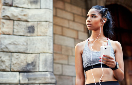 She moves to the beat of her own drum. an attractive young woman listening to music and using her cellphone while exercising outdoors in the city.の写真素材