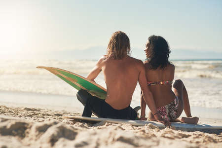 I fell in love with his passion for surfing. a young couple sitting on the beach with their surfboards.の写真素材