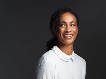 The most beautiful woman is the one smiling. Studio shot of an attractive young woman wearing a white blouse and smiling against a dark background.の写真素材