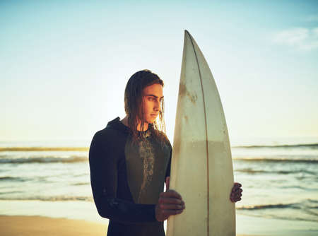 Im out to chase some waves. a young man standing with a surfboard at the beach.の写真素材
