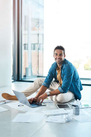 Hard work keeps me ahead in the industry. Portrait of a young businessman brainstorming with a laptop and paperwork on a floor in an office.の写真素材