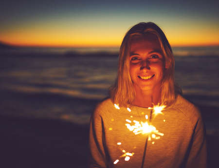 Dont let anyone dim your light from within. Portrait of a young woman holding a sparkler at the beach at night.の写真素材
