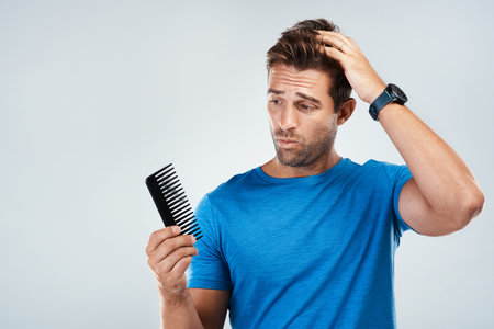 Okay I might have to give it a try. Studio shot of a young man with a confused facial expression holding and looking at a hair comb while standing against a grey background.の写真素材
