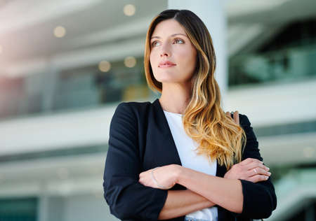 She knows exactly where she wants to be. an attractive young businesswoman looking thoughtful while standing with her arms crossed in a modern office.の写真素材