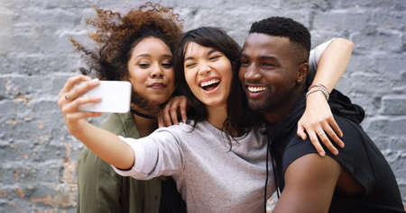 Its selfie time guys, strike a pose. a group of friends taking a selfie together while posing outdoors against a grey brick wall.の写真素材