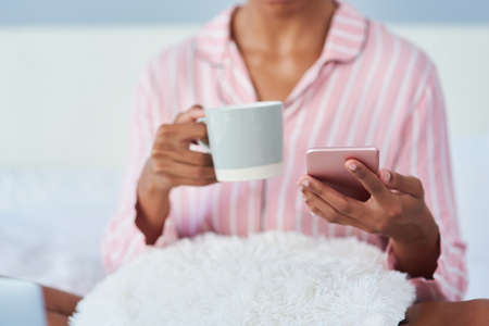Keeping you connected from the start till finish. an unrecognizable woman drinking coffee and using her cellphone in bed at home.の写真素材