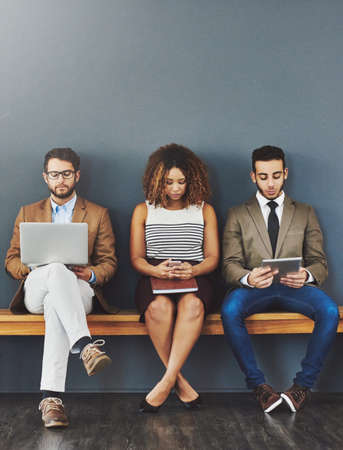 Group of design interns on wireless online devices waiting at a job interview of a startup company. Diverse people sitting, waiting for a meeting with human resources for a jobの写真素材