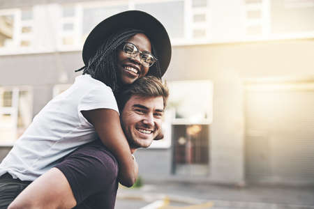 Young playful interracial couple playing, bonding and hugging while standing in the city together on weekend. Happy husband giving wife a piggyback ride, enjoying a day in the urban town or travelingの写真素材