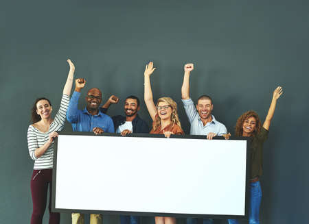 Excited business people showing a blank sign, promoting a product and giving a message on a board while standing together in an office at work. Portrait of happy colleagues holding an empty posterの写真素材