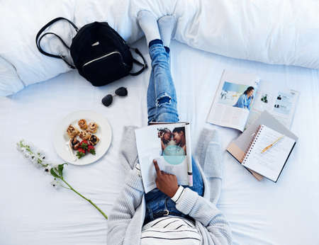 Being a blogger has become her hobby. High angle shot of an unrecognizable woman reading a magazine and having breakfast on her bed in her bedroom at home.の写真素材