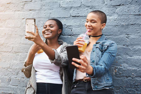Cuteness overload. two young women standing beside a building smiling and taking selfies while holding their cool drinks.の写真素材