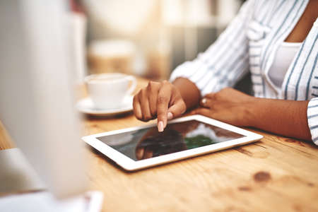 Female african hand using a tablet at a desk doing office work. Closeup of a professional woman browsing on social media while having a cup of tea or coffee. Businesswoman doing research online.の写真素材