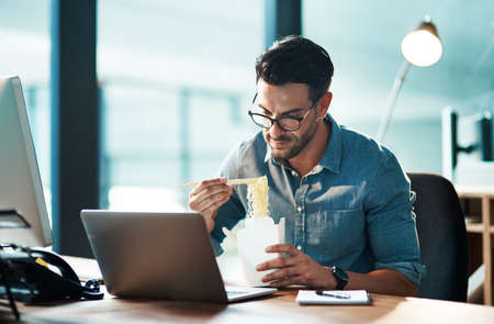 Business man eating lunch at his desk, reading an email on a laptop and working overtime in office. Corporate professional, manager or employee completing a deadline and browsing internet for ideasの写真素材