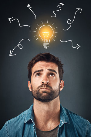 Im confused... Studio shot of a handsome young man looking confused while looking up at an illustration of a light bulb against a grey background.の写真素材