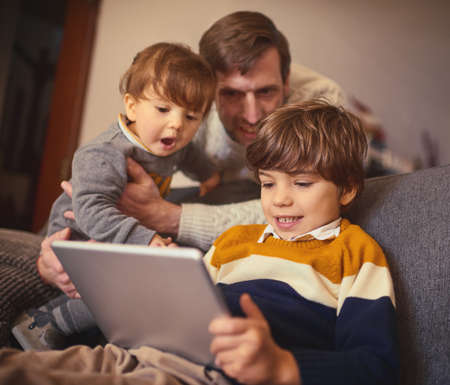 Lets see what your brothers doing. an adorable little boy using a tablet on the sofa at home while his father and younger brother look on.の写真素材